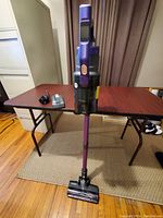 Full view of the purple and gray cordless stick vacuum cleaner standing upright on carpet, showing the main body, electronic handle, and cleaning head.
