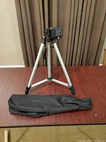 Photo showing the full camera tripod set up on a table with a black carrying case laid flat beside it.