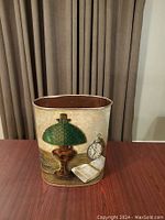 Front view showing decorative design of green lamp, pocket watch, and book on vintage tin waste can.