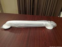 Photo of a white 18-inch grab bar wrapped in plastic packaging, placed on a wooden surface with brown curtains in the background.