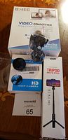 Three brand new electronic devices including video converter box, mini wireless camera with accessories, and phone tripod selfie stick boxed.