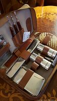 Photo of antique silver grooming set showing various grooming tools, containers, comb, brushes, and razor inside a zippered case with brown leather holders.