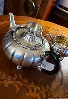Photo showing the antique British silver plate teapot with ribbed design, black handle, and lid with finial, alongside the sugar cup and creamer.