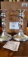 Pair of shiny silver-tone candlesticks standing in front of original cardboard box with handwritten "Silver Candlestick (2)" and a pewter care instruction pamphlet visible on the table.