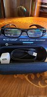 Black eyeglasses with built-in camera resting on top of original box, showing front view and accessories below including USB charging cable and power adapter inside carrying case.