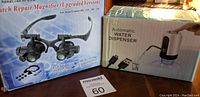 Two boxed items on table: watch repair magnifier glasses with LED lamp, and automatic water dispenser with USB cable shown on box.