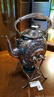 Full view of the antique silver plated teapot on the metal stand with warming base below, showing body, spout, and handle.