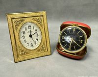 Two vintage travelling alarm clocks shown together, one with a gold-tone ornate frame and white face, the other in a red leather travel case with black face.