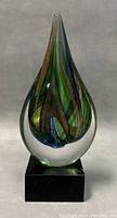 Front view of the colorful blown glass teardrop sculpture on black base, showing internal swirls of green, blue, and gold.
