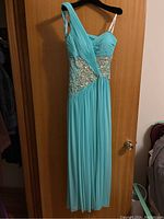 Full view of long turquoise evening gown hanging on door, showing flowing fabric, pleated bodice, and asymmetric straps.