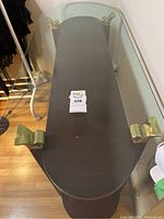 Top view showing oval glass top with four brass brackets and dark wood base underneath.