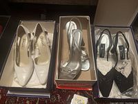 Photo of three pairs of designer women's shoes side by side in their boxes: white Bally pointed toe pumps, silver Dezario slingbacks, black Stuart Weitzman slingbacks.
