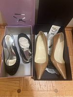 Two pairs of women's high heel shoes in their original boxes on the floor; black slingback X Fashion with cutouts and beige patent Prada pumps side by side