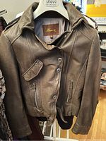 Brown leather bomber jacket on hanger showing front buttons, zipper, flap pocket and zippered pockets