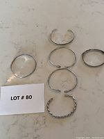 Full view of six silver bracelets on surface, varying styles and textures, with lot number card.