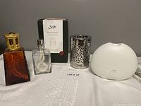 Photo shows four diffusers: two Lampe Berger glass diffusers (one amber with gold-tone top, one clear with silver perforated top) and two Saje diffusers (one white rounded diffuser and one new in box) displayed on white surface.