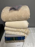 Stack of seven blankets and throws, showing variety of colors and textures including beige plush blanket on top with sealed Sunbeam electric heated throw packaging.
