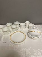 Full set view showing 5 Pyrex mugs, 3 Corning mugs, 5 Butterfly Gold luncheon plates and 5 cereal bowls arranged on tablecloth.