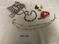 View of the entire lot showing multiple jewelry pieces on a white patterned fabric including pearl necklace, pearl bracelet, Brighton bracelet, red cord charm bracelet, holiday pins, small textured coin purse pouch, and floral drawstring bag.