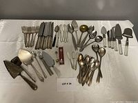 Photo showing a large assortment of mixed cutlery items including forks, knives, spoons, and various serving utensils laid out on a white tablecloth.