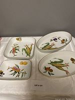 Four porcelain serving pieces with fruit and vegetable patterns and some gold trim, displayed on a white tablecloth