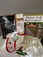 Lot showing Deluxe Golf Ball Cabinet box, cabinet itself wrapped in plastic, white golf bag filled with golf balls and accessories on cloth surface.