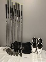 Nine chrome finish irons standing upright beside black Etonic shoe bag and white/black Etonic golf shoes.