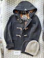 Top down photo of dark grey Aritzia TNA wool hooded coat with toggle buttons and plaid lining, laid flat with two knitted toques underneath (one grey, one cream).