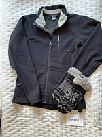 Black MEC jacket and rabbit trimmed gloves laid flat on white quilted surface with LOT #125 card visible