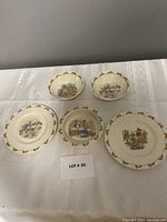Overall view of five Royal Doulton Bunnykins pieces including plates, bowls, and porringer laid out on a white quilted surface.