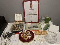 Wide view photo showing all items of the lot on a table including the Eaton's lace tablecloth box, napkin rings on a plate, Pimpernel placemats, glass jar, and various kitchen utensils.
