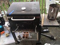 Full view of black Expert Grill BBQ grill with attached side shelf holding a charcoal chimney starter. Grill appears used and dusty, situated outdoors.