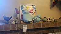 Wide shot of various bird figurines arranged on brick ledge in front of a colorful bird tray and ceramic bird cookie jar.