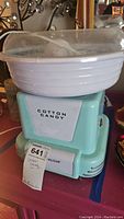 Front view of the Nostalgia cotton candy machine showing the blue base with 'Cotton Candy' label and clear plastic cover on top.