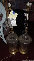 Photo displaying two matching crystal lamps with brass bases and clear crystal finials, no lampshades, placed on a carpeted floor