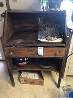 Front view of antique wooden desk showing work surface, drawer with two round knobs, and open lower storage containing various items. Desk surface has stains and wear marks.