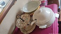 White ceramic covered soup tureen with ladle, two-tiered floral serving plate, yellow and white ceramic dishes on a pink plastic storage bin.