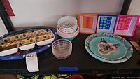 Dishware items displayed on black surface including blue oval platter with white stars, polka dot divided dish, small patterned and clear bowls, cat face plates, and colorful plastic trays.
