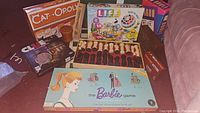 Photo showing several board games including Cat-Opoly, The Game of Life, Clue, The Barbie Game, and Exploding Kittens all stacked together on a carpeted floor.