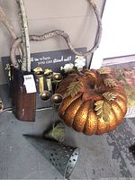 View of all items including large metal pumpkin, cone-shaped metal stand, wooden vase with branches, black metal sign with faux flowers