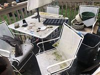 Photo showing four white metal outdoor chairs with mesh seats and backs around a metal patio table. The chairs have dirt and green moss spots from outdoor exposure.