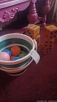Four large wooden orange dice with black dots next to a white and green bucket with bocce balls inside on a maroon carpet.