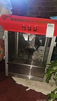 Front view of Roosevelt Popper popcorn machine with red top and visible kettle inside, three switches on front. Glass panels and stainless steel body.