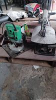 Photo showing two power tools side by side: a green Hitachi TR-12 router and a grey Makita SJ401 bench saw, placed on a surface in a shed environment.
