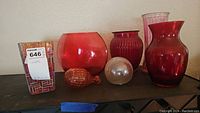Lot shows six glass items positioned on a shelf including a square patterned red vase, round red vase, fish-shaped glass piece, pearl-like glass sphere, ribbed narrow red vase, and a tall clear ribbed vase.