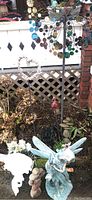 Photo of the outdoor yard art including the resin fairy sculpture, a rock stack next to the fairy, white resin bird garden stake, heart-shaped hanging decoration made of shells and the metal whirligig in the background.