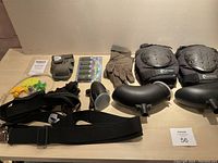 Wide view of laid out paintball accessories including protective gear, loaders, straps, and air hose pieces.
