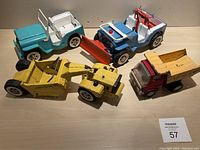 Overhead view of four Tonka toy vehicles on light wood surface: grader, dump truck, jeep and plow jeep.