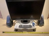 Front view of LG Flatron monitor turned off with Logitech speakers, remote control on stand, and webcam, set on a table with a measuring tape showing about 16 inches wide.