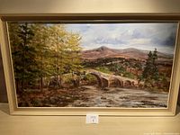 Wide view of the framed painting showing entire artwork with scene of stone bridge, trees, flowing stream, and mountains.